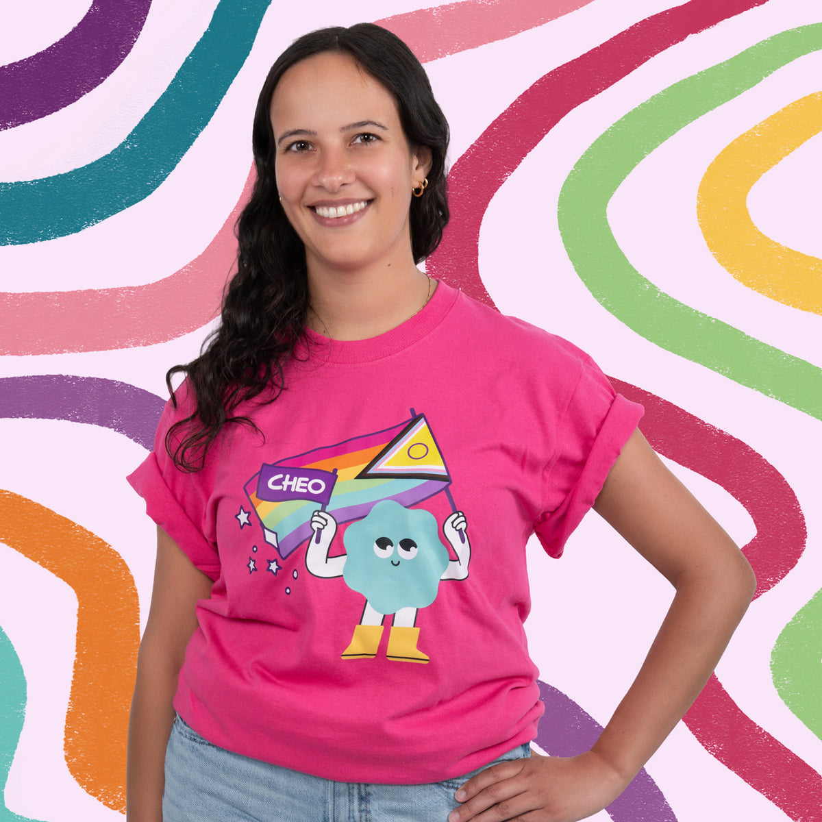 A woman smiling and wearing a bright pink graphic t-shirt with a cartoon character and the word 'CHEO' on the front, set against a colorful wavy background.